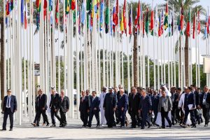 group of people in suits walking beneath limp flags on tall poles